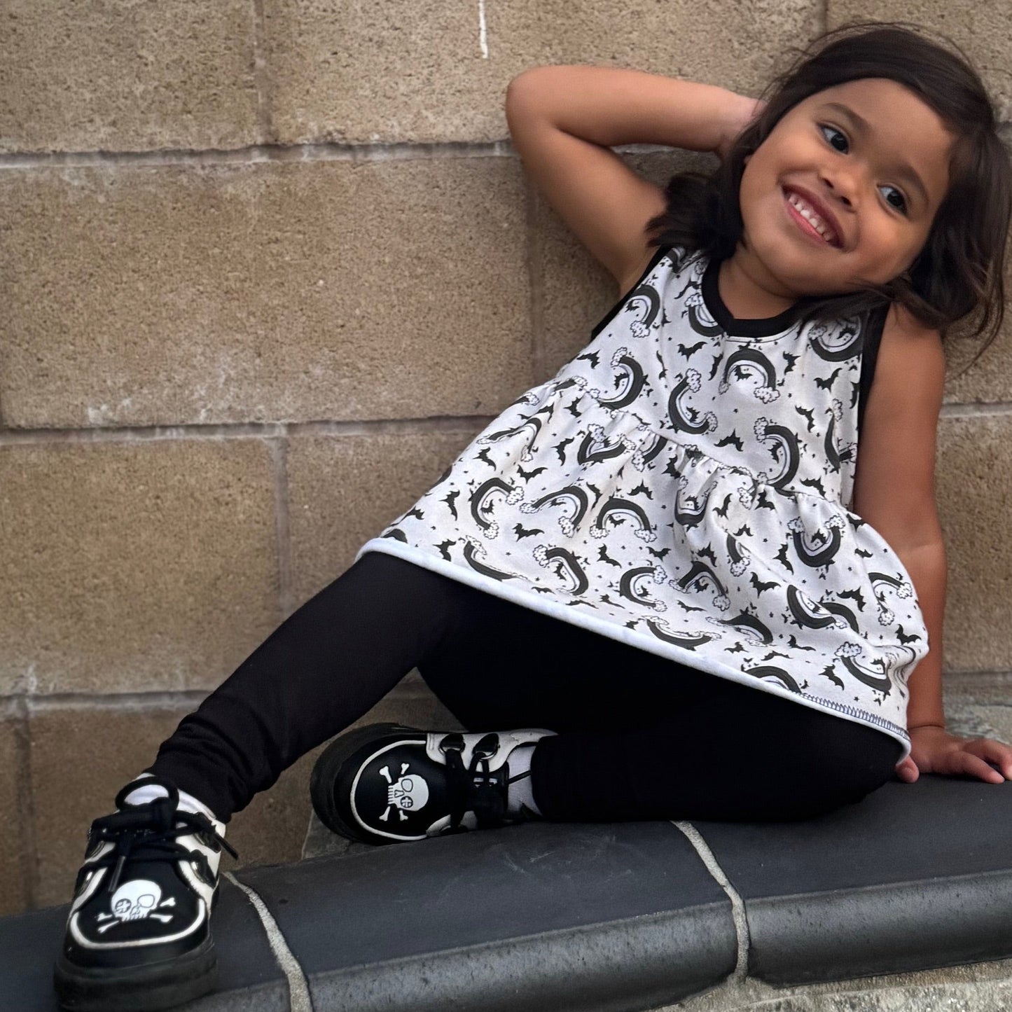 Child wearing alternative clothes : a black and white patterned peplum tank top and shoes with skulls sitting on a ledge against a stone wall.