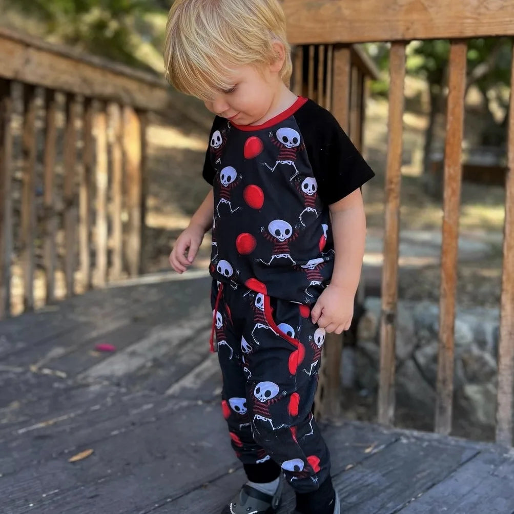 Goth baby boy wearing a matching outfit, black joggers with pockets, raglan top. The pattern features playful skeletons with red balloons.