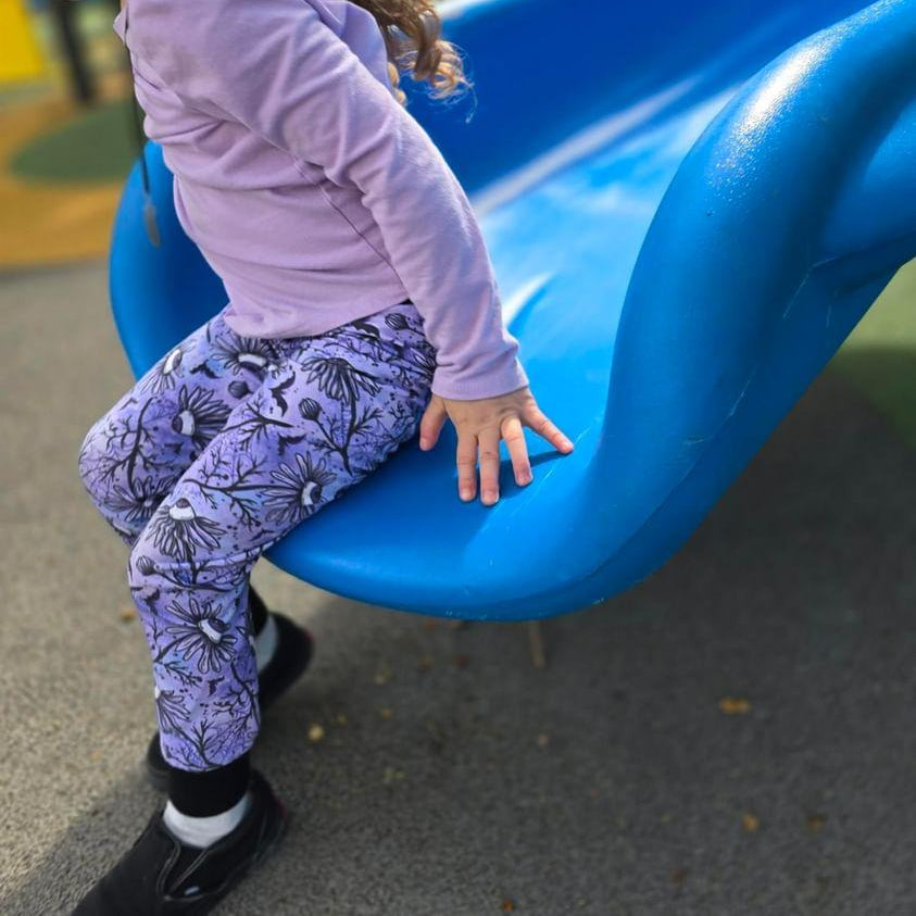 A child wearing spooky floral purple leggings is sitting on a blue plastic slide. Leggings are handmade in the US.