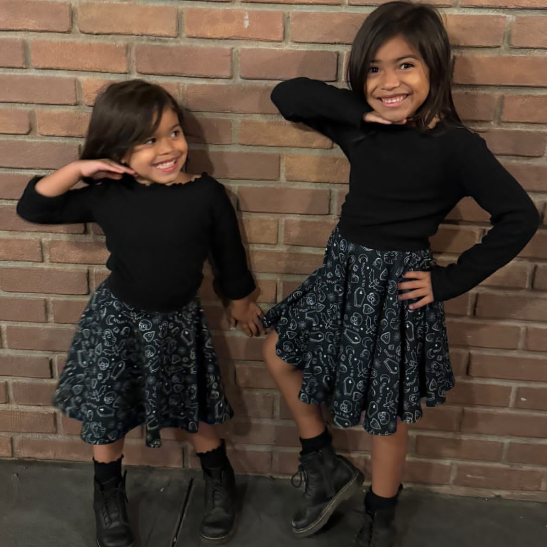 Two children wearing matching outfits in front of a brick wall. Black top, black skirts with halloween - spooky prints