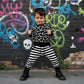 Child wearing a black and white outfit with a bones patterned shirt and striped pants, standing in front of a graffiti-covered wall.