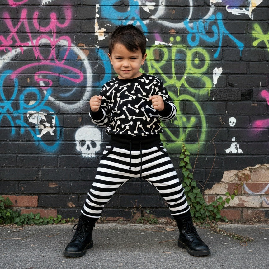 Child wearing a black and white outfit with a bones patterned shirt and striped pants, standing in front of a graffiti-covered wall.