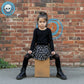 Girl wearing a black sweater with a black skirt with big white bones on it. She's sitting on a cardboard box against a brick wall.