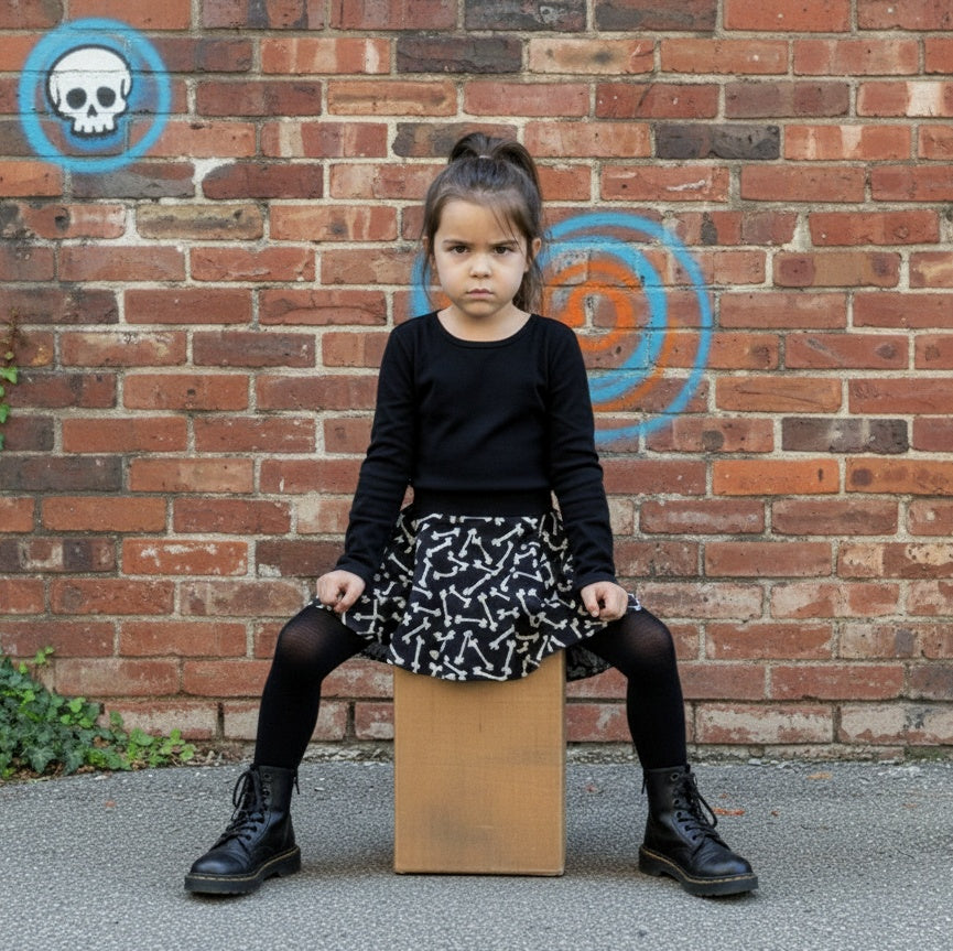 Girl wearing a black sweater with a black skirt with big white bones on it. She's sitting on a cardboard box against a brick wall.