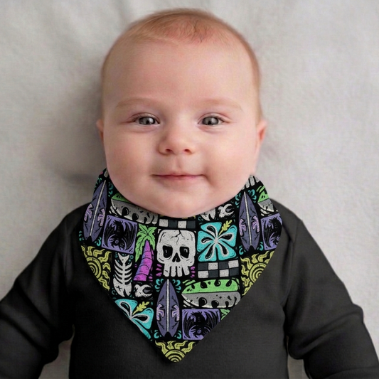 Baby wearing a colorful bandana bib with skull and surfboard design on a neutral background