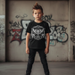 Child wearing a black t-shirt with a white graphic design, standing in front of a graffiti-covered wall.