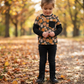 A young child standing on a leaf-covered path in a park, wearing a long-sleeve top with a pumpkin pattern, black pants, and black boots.
