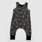 A sleeveless baby romper with an all-over white spiderweb pattern on a black background, featuring a snap-button crotch and black ribbed ankle cuffs, shown against a light gray background.