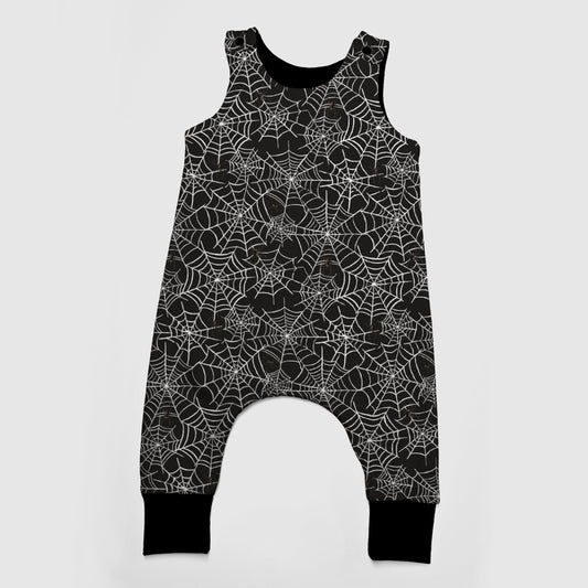 A sleeveless baby romper with an all-over white spiderweb pattern on a black background, featuring a snap-button crotch and black ribbed ankle cuffs, shown against a light gray background.