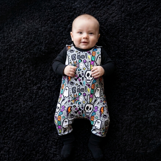A baby wearing a black long-sleeve romper with a colorful Halloween-themed print of ghosts, skulls, and pumpkins, lying on a black textured rug.