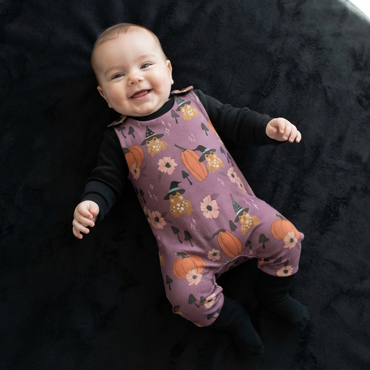 A baby lying on a round black rug, wearing a purple floral and pumpkin print footless romper over a black long-sleeve shirt.