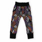 A pair of children's jogger pants featuring a vibrant Halloween-themed pattern with pumpkins, ghosts, spiderwebs, and bats in purple, orange, black, and white, finished with solid black cuffs and waistband.